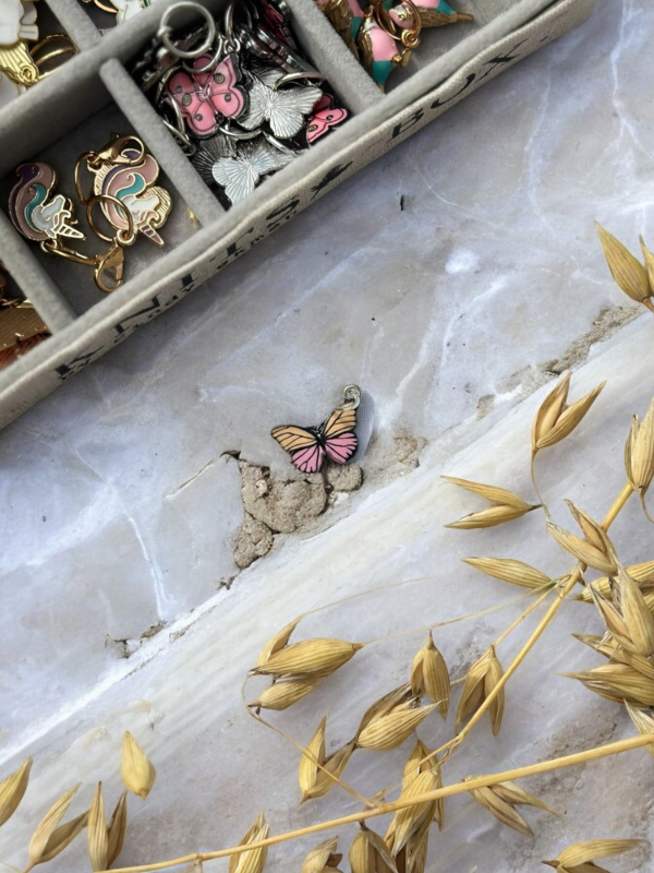 A small pink butterfly charm from KNITS by cindy ekman silmukkamerkit, ideal as kauniit silmukkamerkit for neulonta, rests on a marbled surface beside dried yellow foliage; colorful enamel charms are partially visible in the upper left.