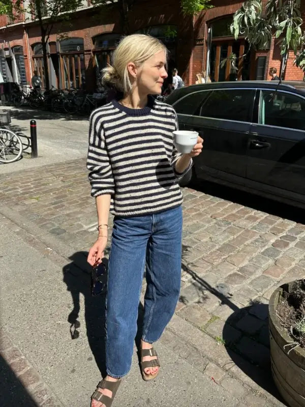 A woman with blonde hair in a bun wears a black and white striped PetiteKnit Olga sweater neuleoohje, blue jeans, and sandals. She holds a white cup and sunglasses while standing on a sunlit cobblestone street beside a parked car.