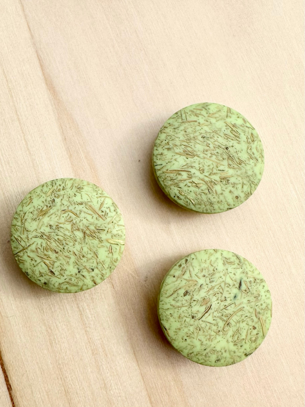 Three round, green Kierrätysnappi kookos 20mm (0454241) buttons with herbal-like flakes rest on light wood, resembling herbal kookosnapit treats.
