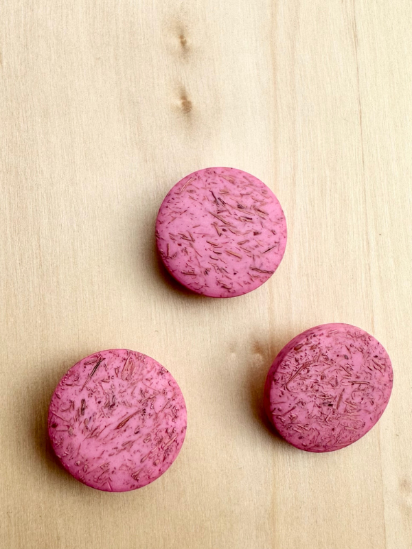 Three round, textured Kierrätysnappi kookos 20mm (0454241) buttons in pink are arranged on a light wooden surface, each with a speckled look and spaced in a loose triangular pattern.