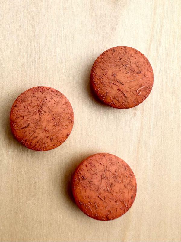 Three round, textured, reddish-brown Kierrätysnappi kookos 20mm (0454241) are arranged evenly on a light wooden surface, displaying a natural and slightly rough appearance.