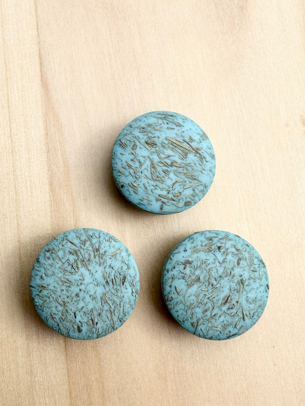 Three round, blue-green Kierrätysnappi kookos 20mm (0454241) buttons with a marbled, textured pattern are arranged in a triangle on a light wooden surface.