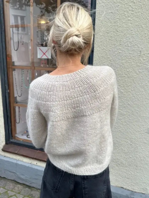 A person with blonde hair in a low bun wears the PetieKnit Anker’s Sweater - My Size neuleohje - Suomi, a light knit top, paired with dark pants, standing outside near a window displaying jewelry.