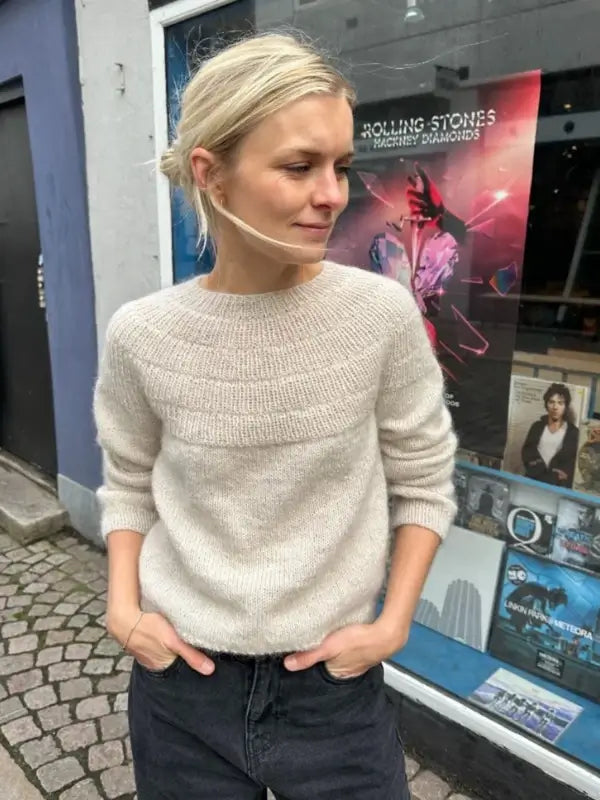 A woman with blonde hair in a loose bun stands outside a shop window, wearing PetieKnit Anker’s Sweater - My Size neuleohje - Suomi—a cozy beige knit paired with dark pants. Her hands are tucked into her pockets, and she wears a transparent face mask under her chin. Posters are visible through the window behind her, and she holds a suomenkielinen printti in one hand.