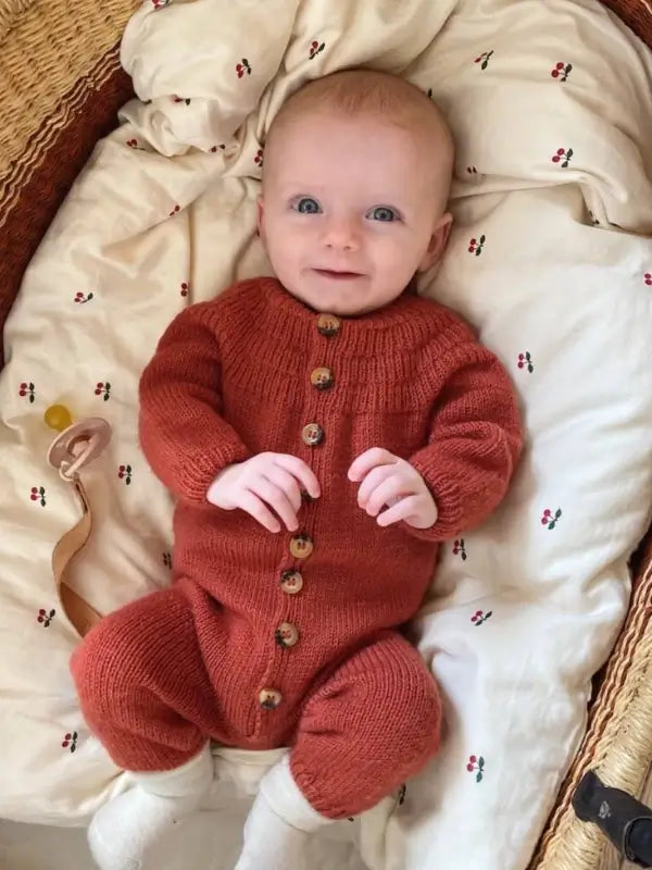 A baby with light skin and blue eyes lies on a patterned blanket, dressed in a rust-colored knit outfit made using the PetitKnit Anker’s Onesie Neuleohje - Suomi pattern, complete with wooden buttons and white socks. A pacifier with a clip rests nearby, creating a cozy scene ideal for a suomenkielinen printti.