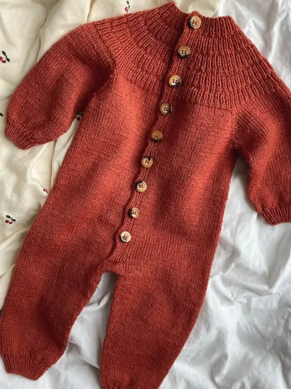 PetitKnit Anker’s Onesie Neuleohje - Suomi: A rust-colored, knitted baby romper with long sleeves and legs, featuring a row of wooden buttons down the front. Inspired by vauvan haalari ohje patterns, the outfit is laid flat on a white fabric background.