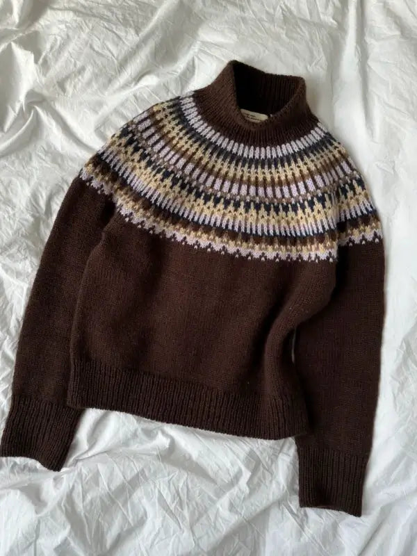 A dark brown PetiteKnit - Celeste Sweater neuleohje featuring a high collar and a circular yoke pattern in white, tan, and yellow tones is laid flat on a crumpled white fabric background.