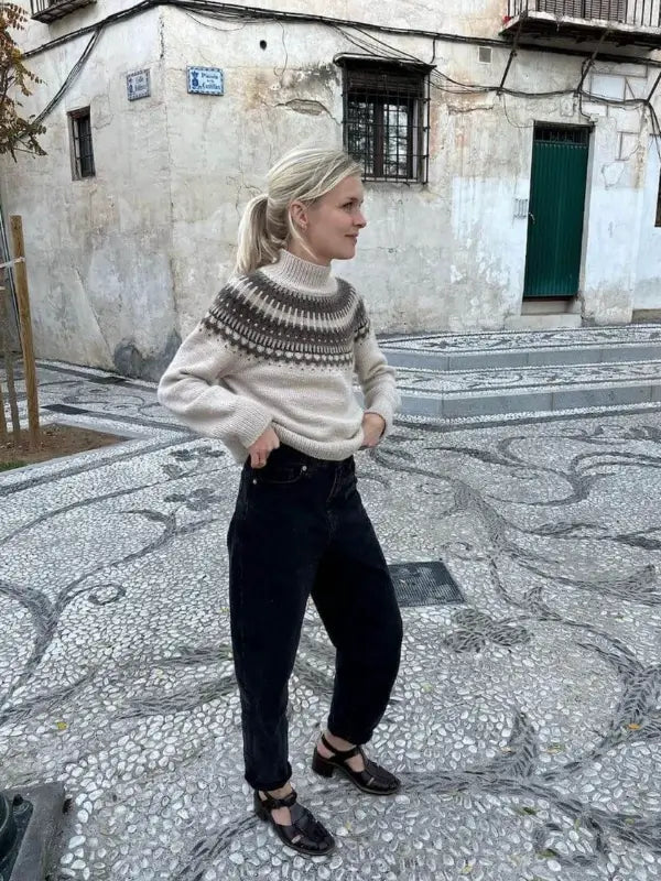 A woman with blonde hair in a ponytail stands on a patterned stone street, wearing the PetiteKnit - Celeste Sweater neuleohje in beige, black pants, and black shoes, in front of an old building with barred windows and a green door.