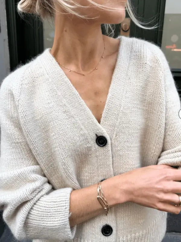 A person wears an oversize, off-white knit cardigan with black buttons inspired by the PetiteKnit - Champagne Cardigan neuleohje, paired with a gold necklace and chain bracelets. The focus is on the upper torso and arms; the face is partially out of frame.
