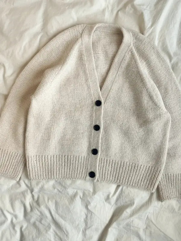 The PetiteKnit Champagne Cardigan neuleohje features a cream-colored oversized design with long sleeves, a deep V-neck, ribbed cuffs and hem, and four dark front buttons—perfect inspiration for your next knitting project.