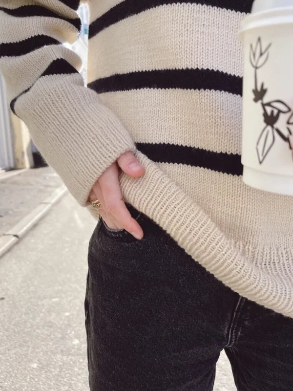 A person wearing the PetiteKnit Marseille Sweater neuleohje- Suomi in beige and black stripes, styled with black jeans, stands outdoors with one hand in their pocket and holding a white cup featuring a black floral design.