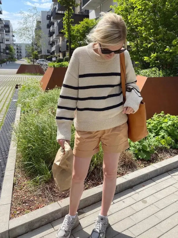 A person wearing sunglasses, tan shorts, sneakers, and the PetiteKnit Marseille Sweater neuleohje- Suomi walks on a sidewalk, carrying a brown bag and an orange tote, with green plants and modern buildings in the background.