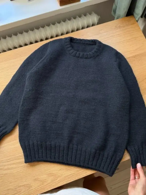 A hand holds the bottom of a dark blue PetiteKnit Niels Sweater Junior neuleohje, Suomi, laid flat on a wooden table near a window with a radiator beneath it.