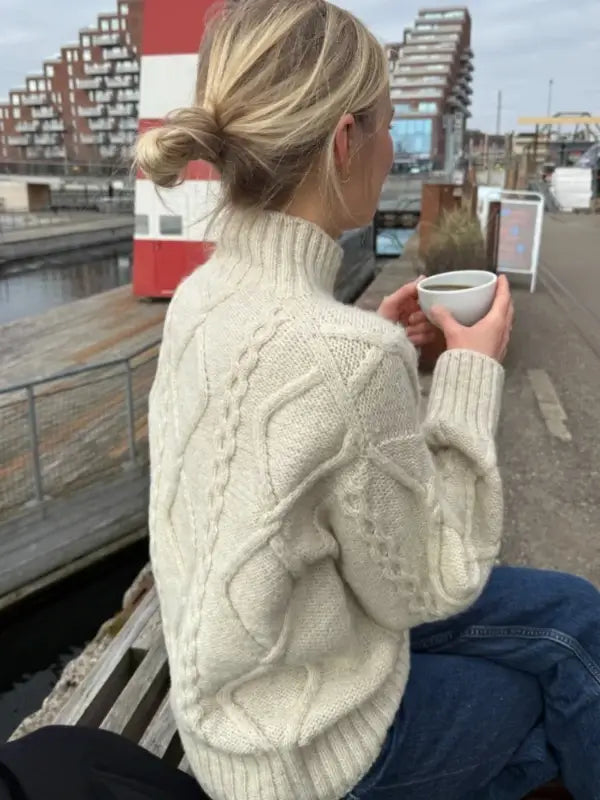 A woman with blonde hair in a low bun, wearing the PetiteKnit Saga sweater neuleohje Suomi in a cream cable-knit style, sits on a bench by the water holding a white mug. Modern buildings and a red-and-white structure are visible in the background.