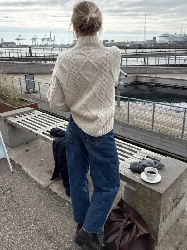 A person wearing a cream cable-knit PetiteKnit Saga sweater neuleohje Suomi and blue jeans stands by the waterfront, facing away. On a nearby bench are a cup of coffee, gloves, and scarf. Industrial cranes and water appear in the background.