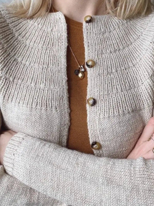 A person with blonde hair wears the PetiteKnit - Anker's Cardigan MY SIZE, a beige knit with a circular yoke and large buttons, over a brown top. Gold necklace and ring complete the look as they stand with arms crossed.