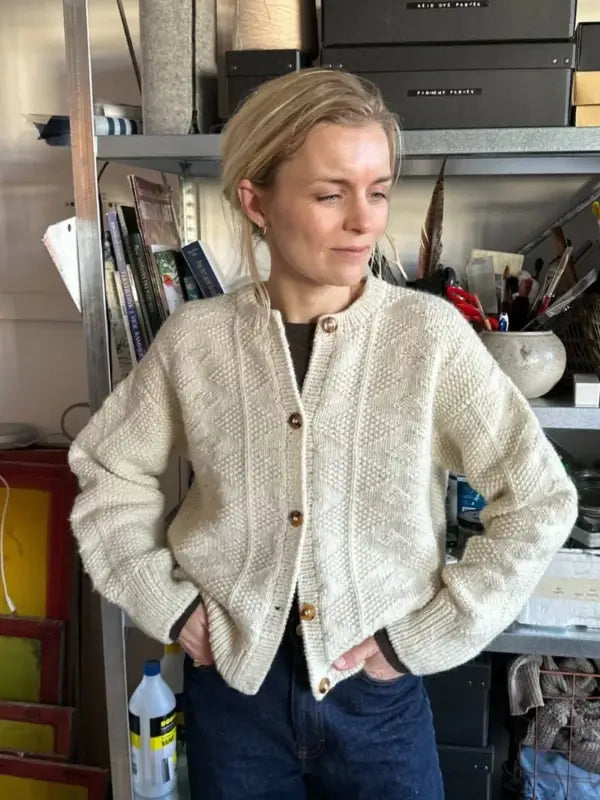 Wearing the PetiteKnit - Esther Jacket Neuleohje, a woman stands before shelves of books and art supplies. The jacket’s cream color, textured knit, and relaxed fit add comfort as she glances to the side with a gentle smile.
