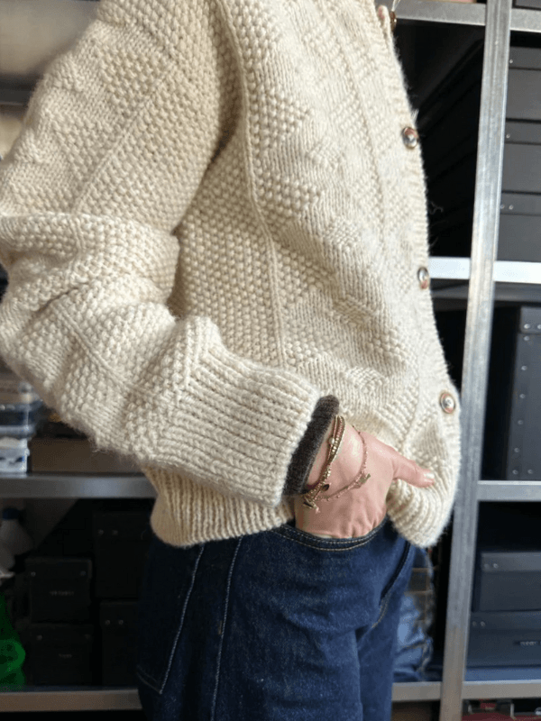A person wearing the PetiteKnit - Esther Jacket Neuleohje, a cream-colored chunky knit sweater with a textured pattern, pairs it with dark blue jeans and stands sideways with one hand in their pocket. Storage boxes are on shelves in the background.