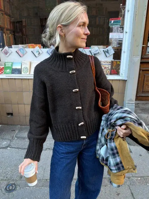 A person wears a dark brown PetiteKnit Hannah Jacket neuleohje -Suomi knit cardigan with a high collar and light-colored toggle buttons.