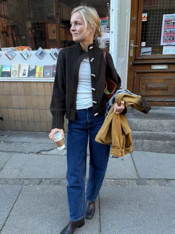 A person wearing a dark brown PetiteKnit Hannah Jacket neuleohje -Suomi with white toggle buttons, a white shirt, and blue jeans, holding a coffee cup and a tan jacket while standing on a sidewalk.