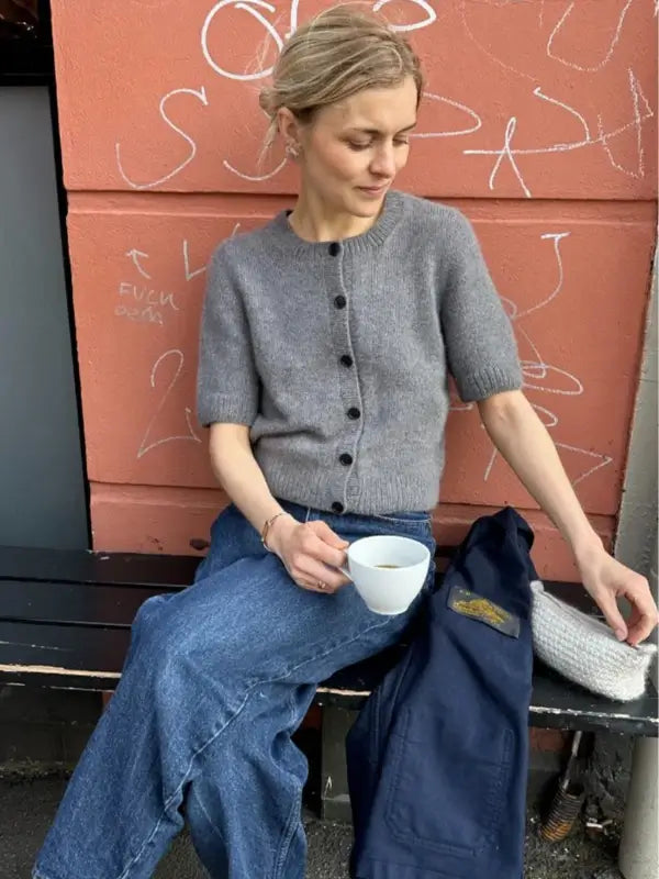 A woman in blue jeans sits on a bench, holding a white cup and wearing the PetiteKnit Maggie Cardigan neuleohje, a buttoned grey short-sleeve style. A jacket and bag are beside her; behind is a pink wall with white graffiti.