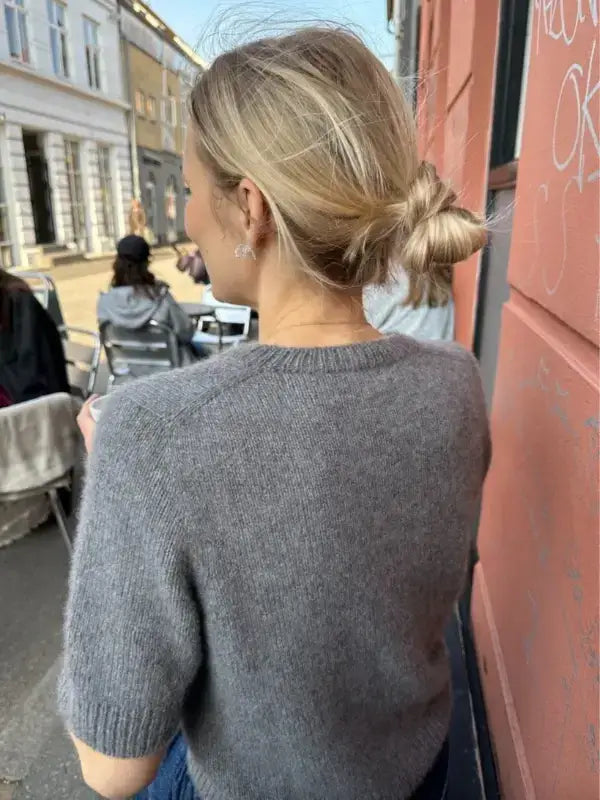 A woman with blonde hair in a messy bun, wearing a gray knit PetiteKnit Maggie Cardigan neuleohje, stands outside near pink and white buildings with people seated at outdoor tables in the background.