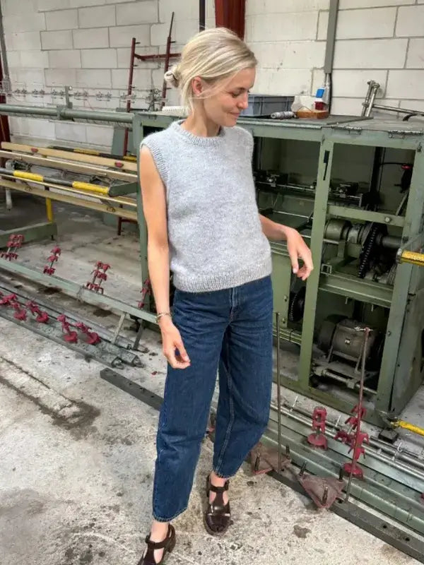 A person wearing the PetiteKnit Novice Slipover neuleohje—a gray, sleeveless sweater—paired with blue jeans and dark shoes, stands indoors near green machinery and metal pipes, looking down with a slight smile.