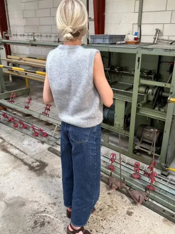 A person with blonde hair in a bun, wearing a gray PetiteKnit Novice Slipover neuleohje and blue jeans, stands indoors facing industrial machinery.