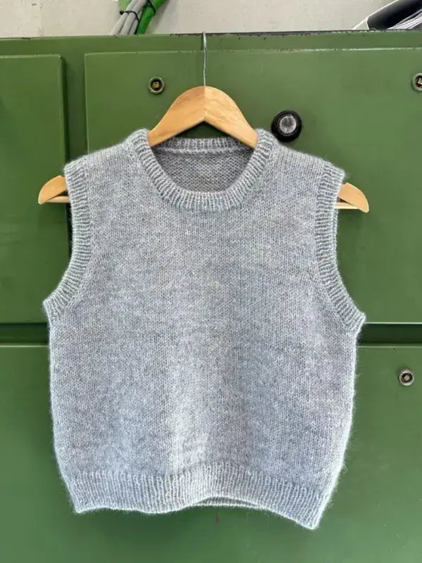 A light gray knitted sleeveless sweater vest, named PetiteKnit Novice Slipover neuleohje, hangs on a wooden hanger against a green background. This stylish neuleohje includes a suomenkielinen printti, making it ideal for knitters of all experience levels.