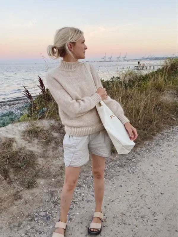 A woman wearing a beige PetiteKnit - Novice Sweater neuleohje, paired with light shorts and sandals, stands on a gravel path by the sea at sunset, holding a white tote bag. Grassy dunes and cranes are visible in the background.