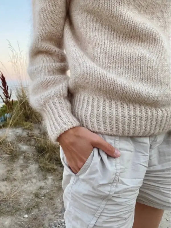 A person wearing a beige knit sweater inspired by the PetiteKnit - Novice Sweater neuleohje stands outdoors with one hand in their pocket. Grassy plants and a hint of sky are visible in the background, evoking the effortless style found in PetiteKnit’s suomenkielinen neuleohje patterns.
