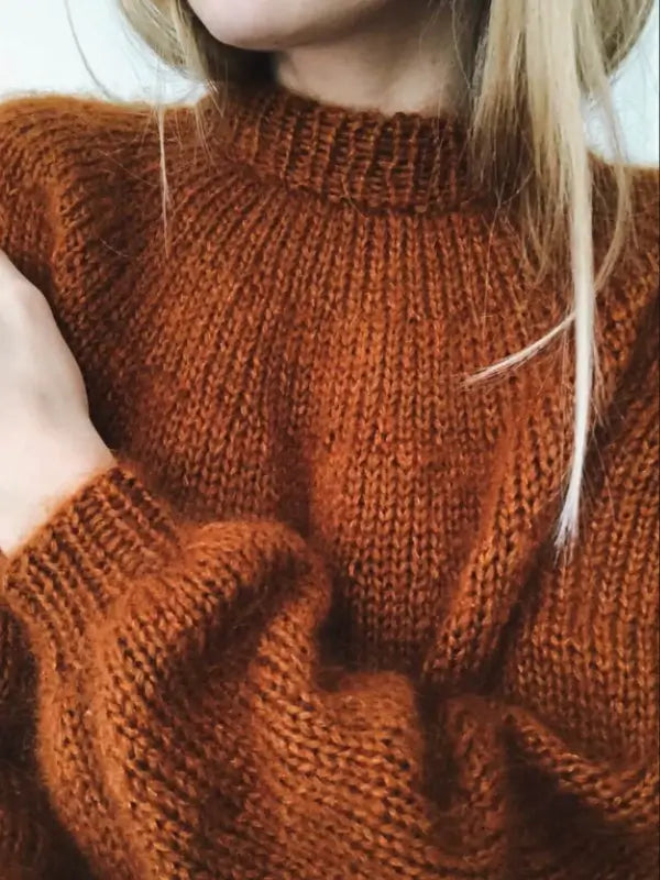 A person with long blonde hair is wearing a thick, cozy, burnt orange knit sweater—possibly handmade using the PetiteKnit -Novice Sweater neuleohje pattern. Only the lower part of their face, neck, and upper torso are visible.