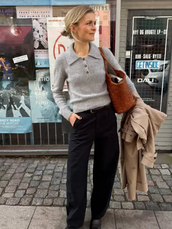 A woman in a gray sweater and black pants stands on a city sidewalk, holding a tan coat and brown woven bag—her style reminiscent of popular designs like the PetiteKnit Petra Polo Neuleohje. Posters are visible on a wall behind her.