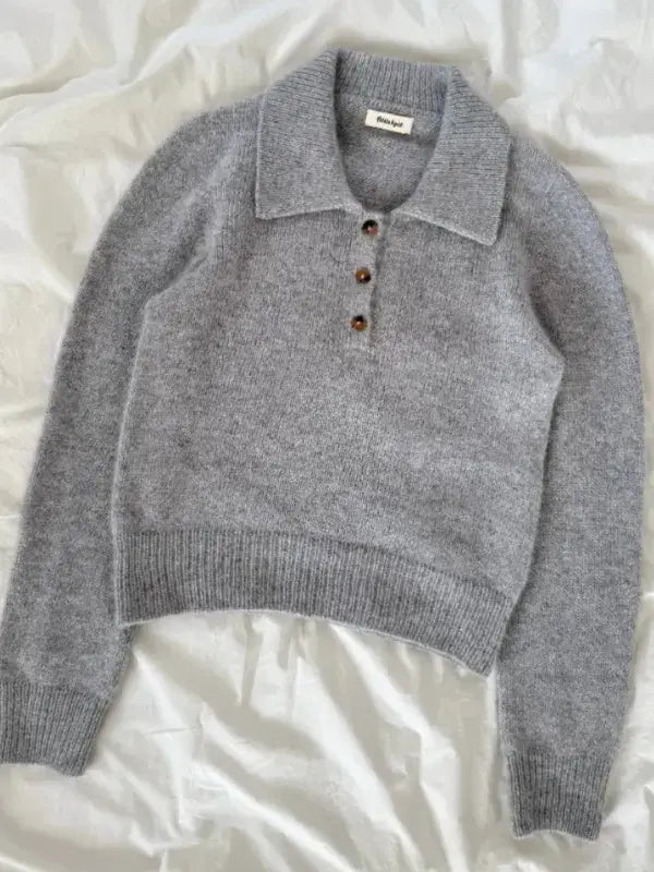 A soft, long-sleeve gray sweater with a wide collar and four brown buttons on the front, laid flat on a white, crumpled fabric background—perfect for knitting with the PetiteKnit Petra Polo Neuleohje or recreating the classic style of the Petra Polo.