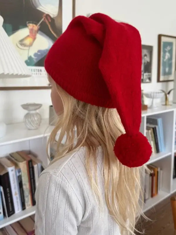 A person with long blonde hair wearing a red Santa-style hat with a pom-pom, possibly made using the PetiteKnit tonttulakki neuleohje, Suomi, stands indoors near a bookshelf, facing away from the camera.