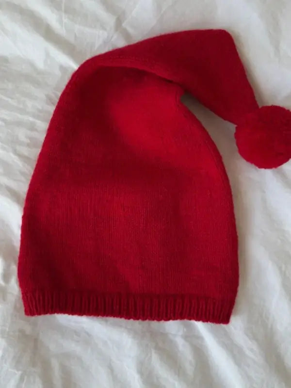 A PetiteKnit tonttulakki neuleohje, Suomi in vibrant red, featuring a pointed tip and a pompom at the end, is displayed on a white textured surface.