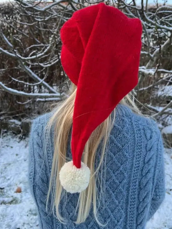 A person with long blonde hair wearing the PetiteKnit tonttulakki neuleohje, Suomi red hat and a blue sweater stands outside in a snowy area with bare branches in the background.