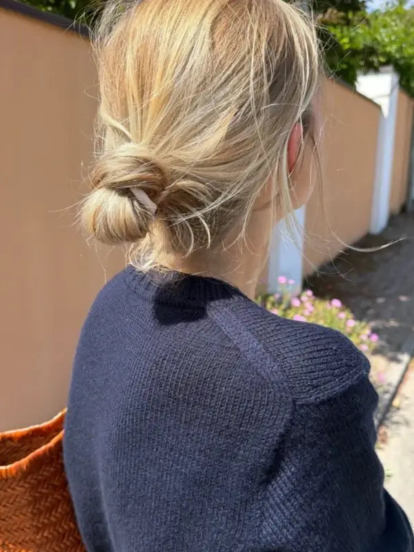 A woman with blonde hair in a loose bun is seen from behind outdoors, wearing a navy blue PetiteKnit Vega Sweater neuleohje and carrying a brown woven bag.