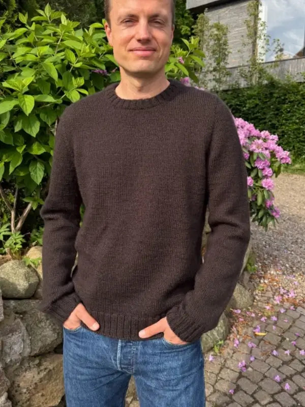 A man stands outdoors with his hands in his jeans pockets, wearing a dark brown Petiteknit Niels Sweater neuleohje, Suomi. Behind him are green bushes, purple flowers, and a stone wall on a sunny day.