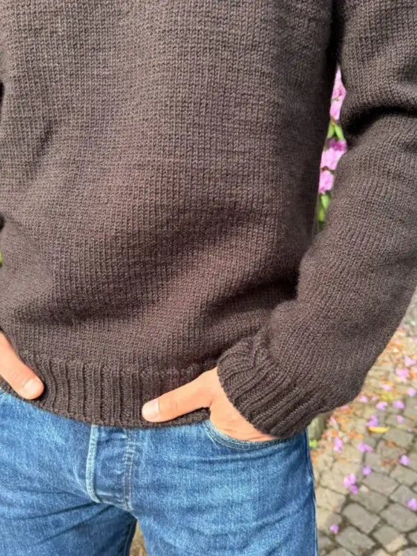A person wearing a dark brown knitted sweater made from the Petiteknit Niels Sweater neuleohje, Suomi, and blue jeans stands outdoors with their hands in their pockets, beside pink flowers and a stone path.