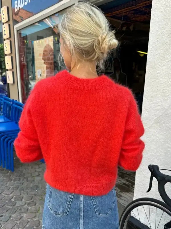A person with blonde hair in a messy bun wears the Petiteknit Stockholm Sweater V-neck neuleohje, Suomi in bright red, artfully crafted from soft silkkimohair, paired with a blue denim skirt. They stand outside near stacked blue chairs and a bicycle, creating a casual and urban scene reminiscent of neuleohje fashion inspiration.