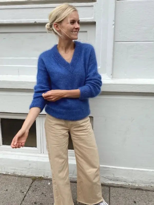 A woman in a blue Petiteknit Stockholm Sweater V-neck neuleohje, Suomi and beige pants stands outside against a white wall, smiling and adjusting her sleeve.