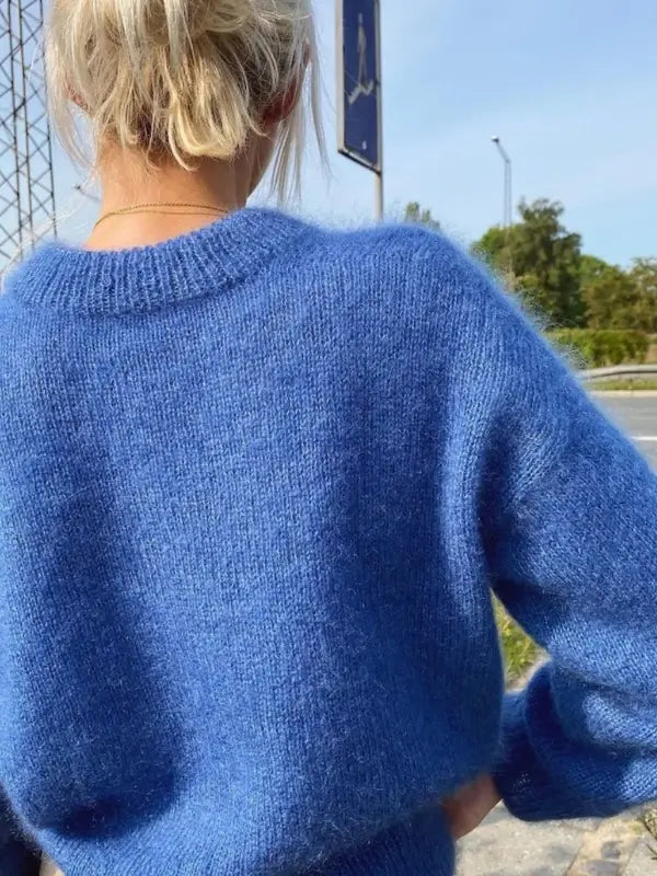 A person with blond hair in a messy bun is seen from behind, wearing the Petiteknit Stockholm Sweater V-neck neuleohje, Suomi and standing outdoors near a road with green trees in the background.