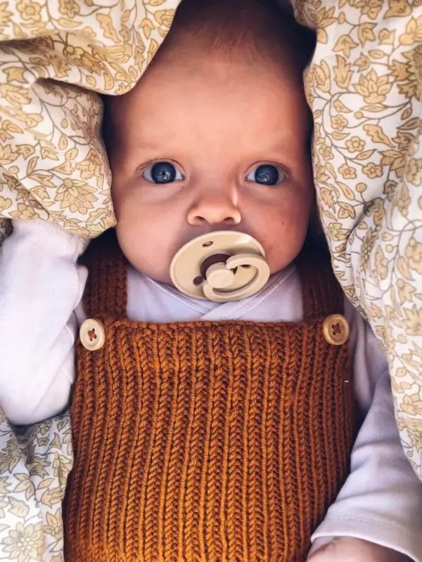A baby with wide blue eyes and a pacifier lies under a beige and yellow patterned blanket, wearing a white shirt and the PetiteKnit Willum’s Overalls neuleohje - Suomi, a brown knitted overall with large buttons.