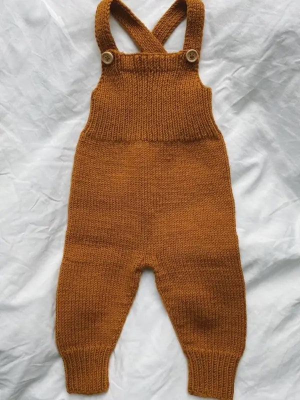 PetiteKnit Willum’s Overalls neuleohje - Suomi features charming brown knitted baby overalls with ribbed cuffs and crossover straps, fastened by two light-colored buttons. Pictured laid flat on a crumpled white fabric background, this delightful haalari is perfect for little ones and makes an excellent knitting project if you’re looking for a new neuleohje.