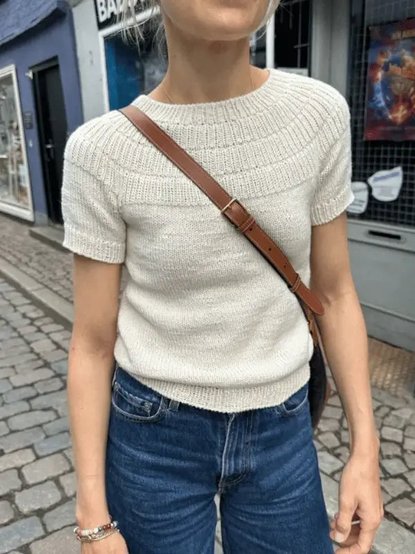 Wearing a cream PetiteKnit Anker's Summer Shirt in Sandnes Garn Line and blue jeans, a person with a brown crossbody bag stands on a cobblestone street, their face partially out of frame.