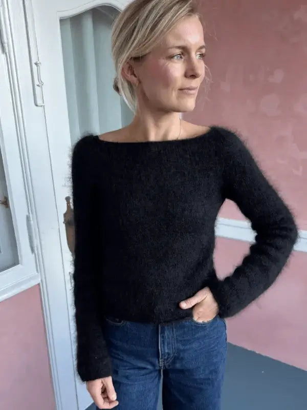 A woman with blonde hair in a loose updo wears a fuzzy black PetiteKnit Bea Blouse neuleohje sweater and blue jeans, standing indoors by a pink wall and white door, looking to the side and smiling softly.