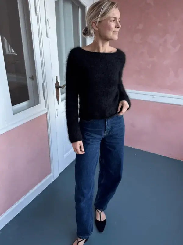 A woman stands indoors wearing a fuzzy black PetiteKnit Bea Blouse sweater knitted with Isager Silk Mohair, paired with blue jeans and black pointed flats. She is looking to the side with one hand in her pocket, in a room with pink walls and a gray floor.