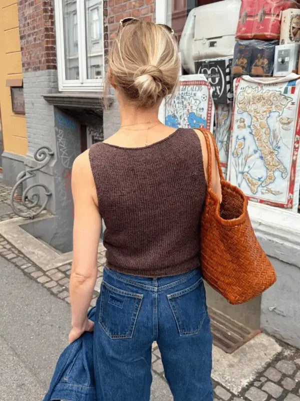 A woman with blonde hair in a low bun, wearing blue jeans and the PetiteKnit Bea Top neuleohje (ENG), carries a large woven brown bag as she stands on a city sidewalk near a wall decorated with posters.