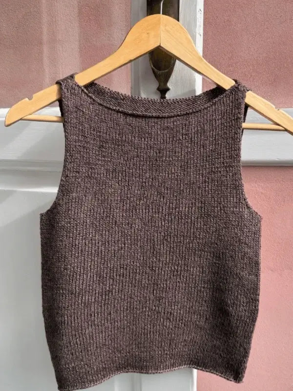 A sleeveless brown tank top knitted from the PetiteKnit Bea Top neuleohje (ENG) hangs on a wooden hanger against a white and pink background.